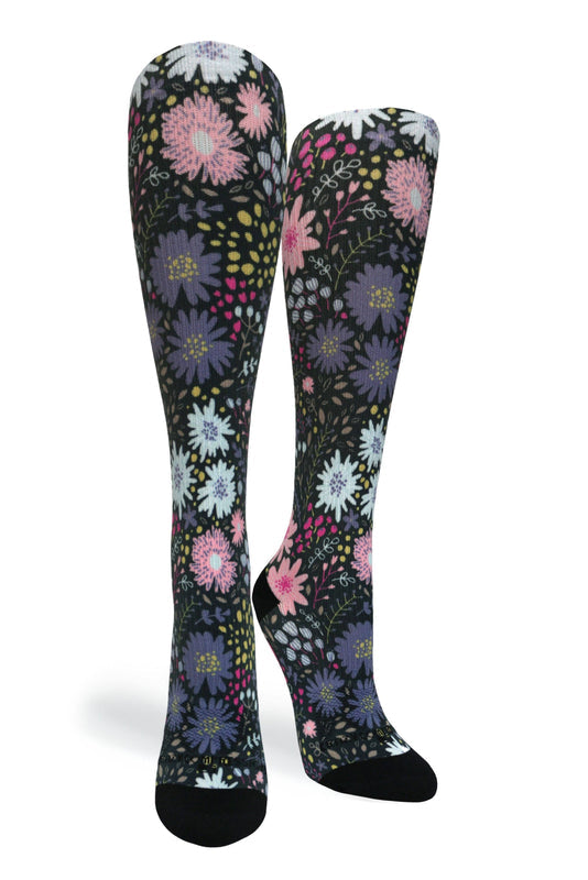 A pair of knee-high socks with a dense floral pattern on a black background, featuring pink, purple, white, and yellow flowers with black toes and heels, shown against a plain white background.